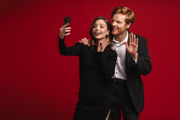 Beautiful couple taking selfie and waving hands while standing isolated over red background