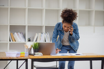Young African American businesswoman using a smart phone and working with pile of documents at office workplace, business finance and accounting concepts.