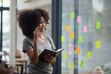Young African American stylish woman working on project plan using sticky papers notes on glass...