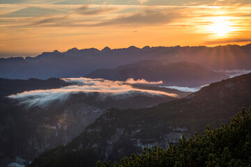 View of an early sunrise during summer as seen from the viewpoint 5 fingers on the Krippenstein mountain toward the valley with fog coming over the mountains and mountain ranges in the background.