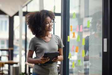 Young African American stylish woman working on project plan using sticky papers notes on glass...