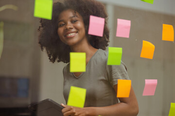 Young African American stylish woman working on project plan using sticky papers notes on glass wall, people meeting to share idea, positive thinking workshop and business design planning concepts.