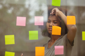 Young African American stylish woman working on project plan using sticky papers notes on glass wall, people meeting to share idea, positive thinking workshop and business design planning concepts.