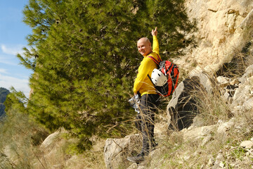 Fototapeta premium mature man, tourist in yellow jacket with orange backpack safety helmet, climber stands front limestone rock, extreme lifestyle, mountaineering, life insurance, difficulties in hike, extreme sports