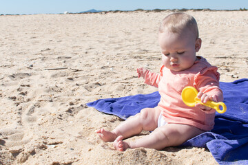 Baby girl playing in the sand with plastic toy. 8 months old baby with blonde hair at the beach.