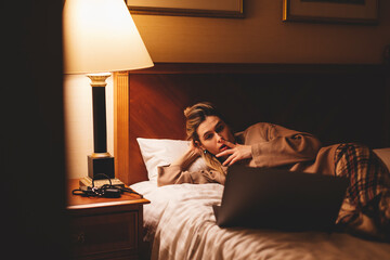 Amazed female watching horror movie on laptop while lying in bed in dark room at night. Afraid...