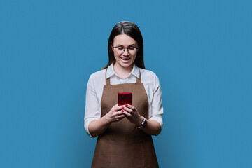 Young smiling woman in apron looking at smartphone, on blue background