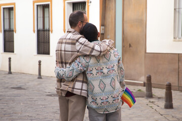 Real marriage of gay couple, hugging on the street, with a gay pride flag in their pocket and seen from the back. Concept lgtb, lgtbiq+, couples, in love, pride, hugging.