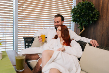 Portrait of attractive young couple in love wearing white bathrobes drinking fresh fruit cocktails, talking, hugging relaxing sitting on comfortable sofa in spa salon by window after beauty treatment