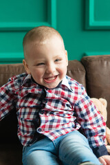 Adorable toddler baby boy in a plaid shirt sits on a brown sofa, looking at the camera and laughing. Big smile, milk teeth.