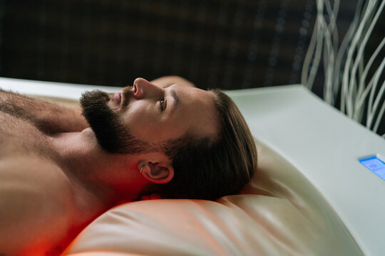 Closeup face of handsome young man lying relaxing on electric massage bed and enjoying spa treatment at luxury spa salon. Muscular male having rest enjoying wellness weekend, thinking looking up.