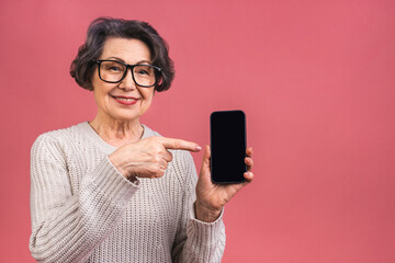 Surprised smiling happy senior mature aged senior woman in casual showing blank smartphone screen while looking at the camera isolated over pink background. Using phone.