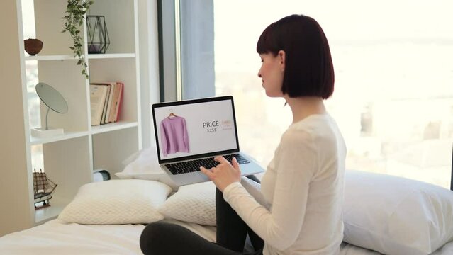 Back view of darked haired woman sitting on white bed and browsing website with trendy clothes on sale. Caucasian female using wireless laptop for choosing new wear with discount of 25 percent online.