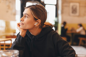 A smiling young woman is sitting in a restaurant and putting wireless earphones on. A woman is relaxing during her coffee break. A young woman in restaurant using technologies listening music.
