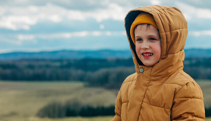 Little boy in yellow jacket and hat stands at outdoor with Sudetes mountains on background © Masson