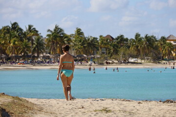 Woman in swimsuit walking by the sand on palm trees background. Ocean tropical beach with tourist resort on Caribbean islands