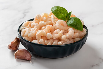 Bowl of frozen cooked shrimps over marble countertop. Peeled orange prawn tails and garlic prepared...