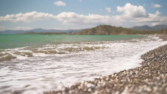 View of the bay on a pebble beach in the Aegean Sea, 4k live video, legs of a man in the frame passing to the sea, selective focus, beautiful sea waves washing the beach, vacation time