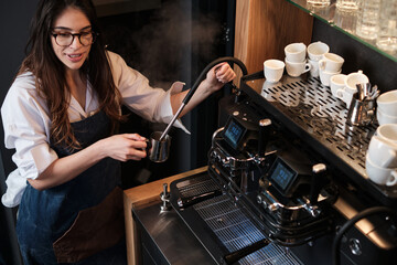Barista standing next to a coffee machine in a coffee shop and heating up milk.