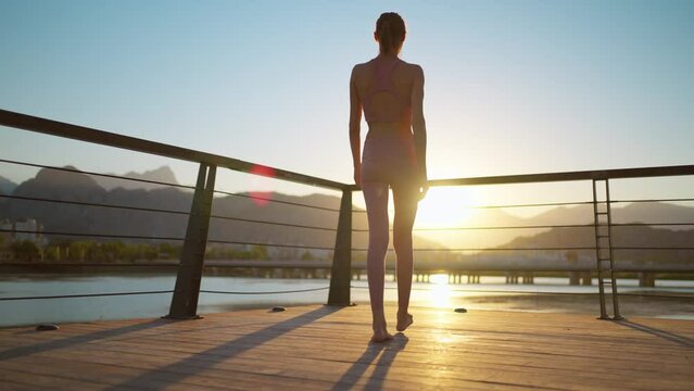 back view of woman silhouette walking barefoot on wooden jetty pier and watching sunset with mountains and sea view. woman relaxing after evening workout