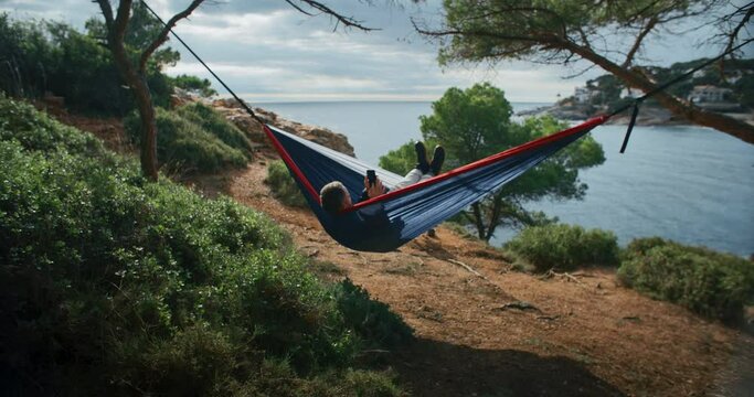 Man chill and relax in camping hammock between two trees in forest on beach shoreline. Relaxing summer or spring vacation. Summertime holiday. scroll through social media on smartphone