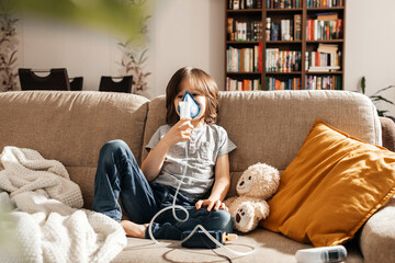A little boy makes an inhalation at home sitting on the couch using a nebulizer and a mask....