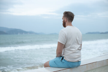 Fototapeta na wymiar Young calm sad serious man is sitting on embankment near sea, ocean on beach, thinking, meditate on natural background 