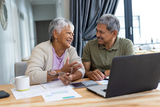 Biracial smiling senior couple with bills and laptop on table discussing finances at home