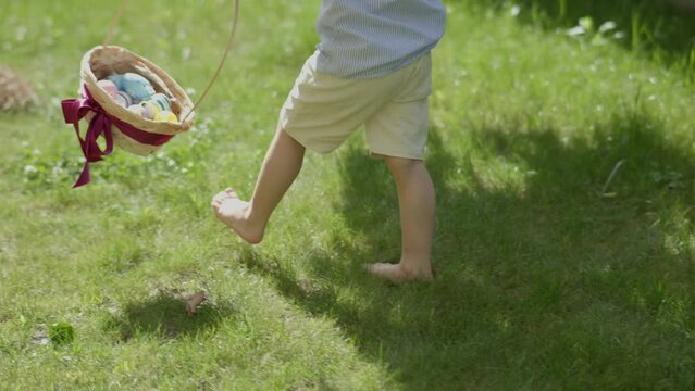child's barefoot legs stepping green grass colorful Easter eggs in basket. traditional spring holiday celebration: little kid boy holding basket full of Easter eggs walking barefoot green lawn