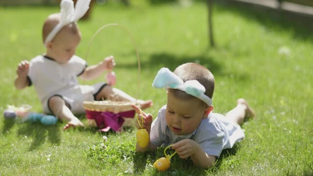 two Easter boys playing Easter eggs on green grass outdoors. Little boy kid bunny ears lying on grass hanging Easter egg on his finger. his toddler brother sit behind wear funny ears. Easter picnic