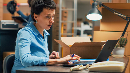 Fototapeta premium Woman entrepreneur looking at merchandise in boxes to plan products shipment at warehouse startup. Female adult using stock to do quality control and work on distribution management. Handheld shot.