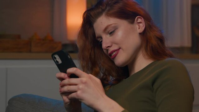 Happy relaxed caucasian redhead woman at home on sofa chatting in social network on phone looking at smartphone screen laughing smiling girl enjoying mobile shopping app in internet using cellphone