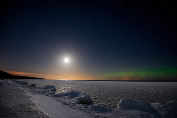 Seashore covered with ice under a starry sky with moon and northern lights. 