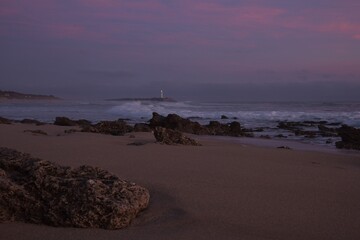 Pink hour view of the clouds above the ocean around Faro de Trafalgar lighthouse seen from Mangueta Beach, Spain, framed by rough dark gray beach rocks and wild waves rolling into Zahora Beach