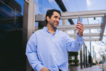 Middle-aged Latin man in an oversized shirt making a video call on his smartphone in front of his office.