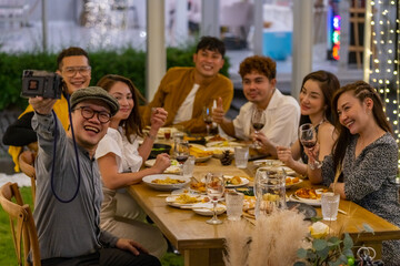 Fototapeta na wymiar Group of Asian people using mobile phone taking selfie together during outdoor celebration dinner party in the garden on summer holiday vacation. Man and woman friend reunion meeting at restaurant.