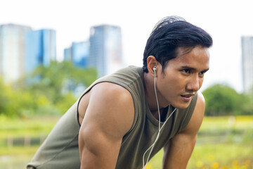 Asian man in sportswear listening to music from earphones with mobile phone during jogging exercise at public park in summer morning. Healthy guy do sport training running workout in the city.