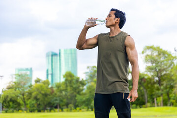 Asian man in sportswear drinking water from a bottle during jogging exercise at public park in the...