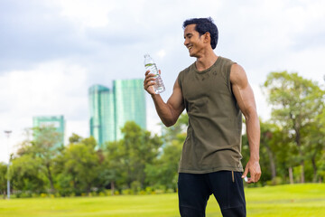 Asian man in sportswear drinking water from a bottle during jogging exercise at public park in the...