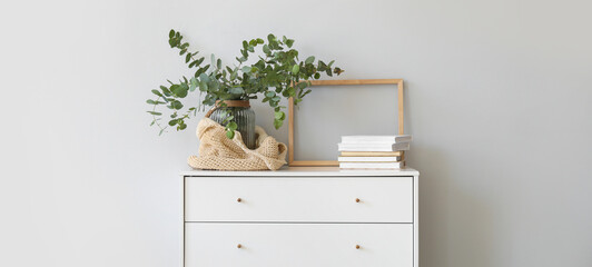Vase with eucalyptus branches, eco bag and books on chest of drawers near light wall