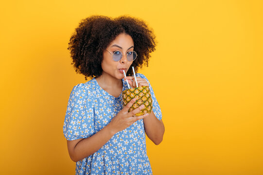 Lovely african american curly woman with blue glasses, in blue summer dress, holds a pineapple in hands, drinks fresh healthy pineapple smoothie, fresh, looks at the camera on isolated pink background