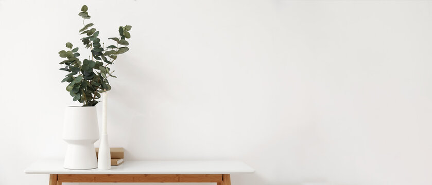 Vase with eucalyptus branches, burning candle and books on table near light wall. Banner for design