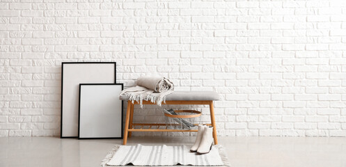 Soft bench with plaid, basket, female shoes and blank frames near white brick wall in room