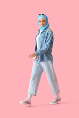 Stylish Muslim woman in blue hijab walking on pink background