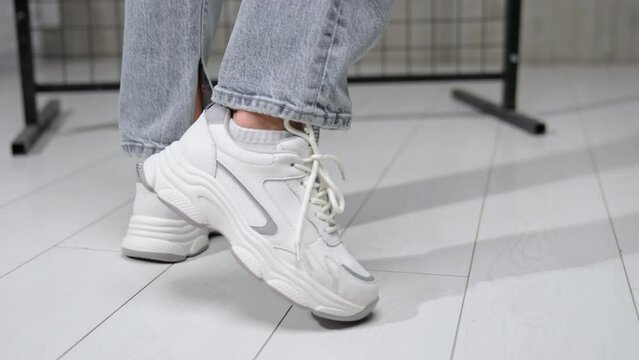 Fashionable white modern sneakers. Female wearing grey jeans walks in comfortable sport shoes by the studio.