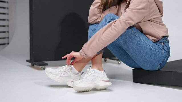 Cropped image of a girl sitting on a black step. Model in jeans, sweatshirt and white sneakers in studio.