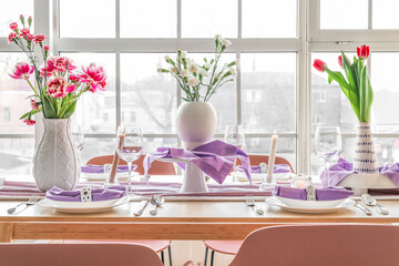 Beautiful table setting for International Women's Day celebration with different flowers and burning candles in light room near window