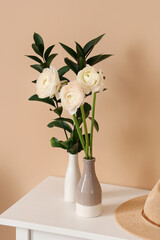 Vases with ranunculus flowers and plant branches on table near beige wall