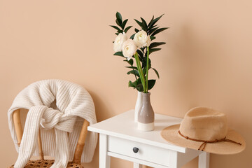Vases with ranunculus flowers, plant branches and hat on table near beige wall in room
