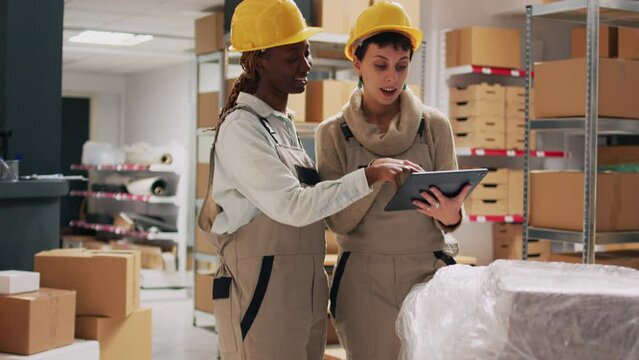 Two women with hardhats organizing products on shelves, looking at tablet to check stock logistics and planning distribution. Warehouse manager and employee counting cardboard packs.